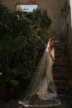 a woman in a wedding dress and veil standing on some stairs with greenery behind her