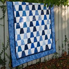 a blue and white quilt is hanging on a fence next to a green tree with leaves