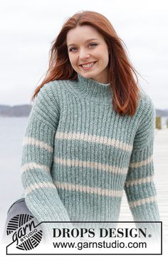 a woman standing on a dock wearing a green sweater and smiling at the camera with her hands in her pockets