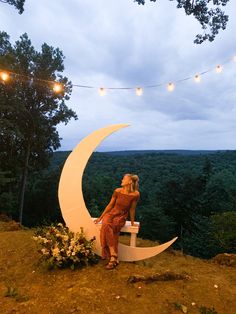 a woman sitting on top of a bench next to a giant crescent shaped moon in the middle of a forest