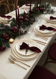 the table is set with white plates and red napkins, silverware, pine cones, evergreen branches, candles, and burgundy ribbon