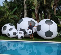 a man standing next to three giant soccer balls near a swimming pool with palm trees in the background