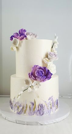 a three tiered cake with purple flowers on the side and white icing around it