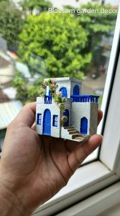 a hand holding up a miniature house made out of clay and painted white with blue trim