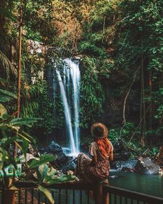 a woman standing in front of a waterfall with her back to the camera and looking at it