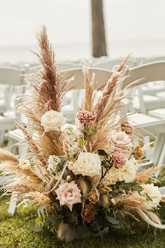 an arrangement of flowers and grasses sits on the grass in front of white folding chairs