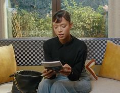 a woman sitting on a couch reading a book and looking at her phone while holding a purse