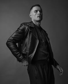 a man in a black leather jacket and slacks poses for the camera with his hands on his hips