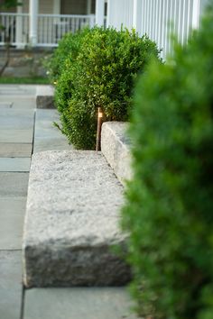 a stone bench sitting next to a lush green bush in front of a white house