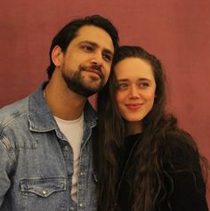 a man standing next to a woman in front of a red wall and smiling at the camera