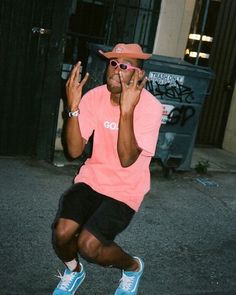 a man in pink shirt and black shorts holding up his hands to his face while sitting on a skateboard