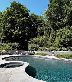 an outdoor swimming pool surrounded by greenery and stone patio furniture, with trees in the background