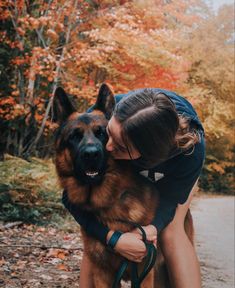 a woman hugging her german shepherd dog in the fall leaves with trees and foliage behind her