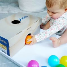 a baby playing with an orange in a box on the floor next to colorful balls