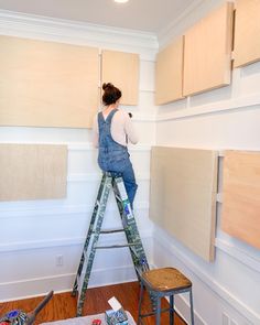 a woman standing on a ladder painting the walls in her home with white paint and wood paneling