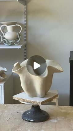 a white vase sitting on top of a wooden table next to shelves filled with pottery