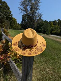 This tumeric yellow hand burnt wide brim fedora would make a perfect addition to any nature lover's wardrobe. The faux suede earthen yellow material burns a rich brown allowing the design to stand out. On the backside brim of the hat I hand burnt and shaded dandelion plants. On the front of the crown I burnt a single dandelion plant. I distressed the hat with stitch marks along the crown and brim of the hat. The design is original and completely free-handed. For a finishing touch I added a brown Rustic Short Brim Hat For Spring, Vintage Yellow Adjustable Hat, Yellow Western Hat With Curved Brim, Yellow Western Hat With Short Brim, Rustic Brimmed Fedora For Spring, Rustic Festival Hats For Spring, Yellow Brimmed Straw Hat, Rustic Fedora For Spring Festival, Rustic Wide Brim Fedora For Spring