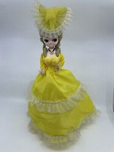 a doll in a yellow dress and hat next to a cell phone on a white surface