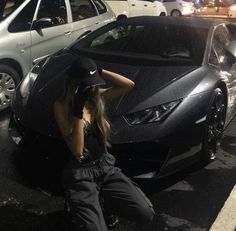 a woman sitting on the ground in front of a black sports car with her hat up