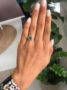 a woman's hand holding an emerald ring on her left hand, in front of a potted plant