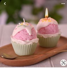 two cupcakes with pink frosting and a lit candle are on a wooden tray