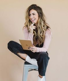 a woman sitting on top of a stool holding a laptop computer in her right hand