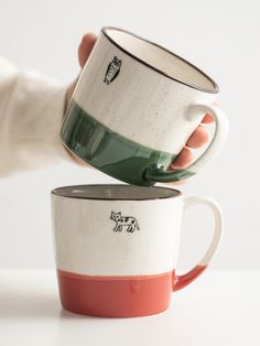 two coffee mugs being held by someone's hand on a white table top