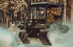 a man riding on the back of a green vehicle through snow covered forest with trees in the background