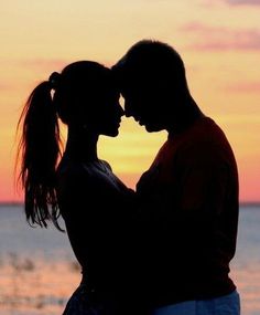 a man and woman standing next to each other in front of the ocean at sunset