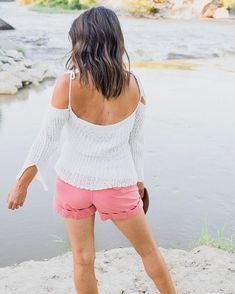 a woman in pink shorts and white top walking on sand near water with her back to the camera