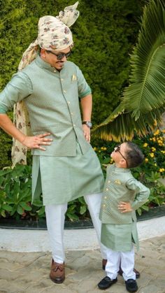 a man standing next to a little boy wearing a green shirt and white pants with his hands on his hips