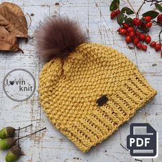 a knitted hat with a pom - pom sits on top of leaves and berries