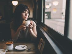 a woman sitting at a table with a cup in her hand and looking out the window