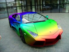 a rainbow colored sports car parked in front of a glass building on a brick sidewalk