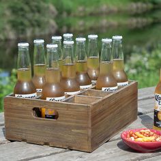 a wooden box filled with bottles of beer next to a plate of food on a picnic table