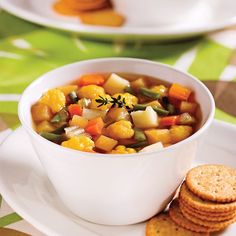 a white bowl filled with vegetable soup next to crackers on a green and white tablecloth