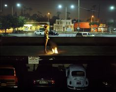 a woman standing in the middle of a parking lot at night with cars parked nearby