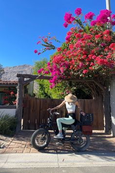 a woman sitting on top of a black bike next to a pink flowered tree