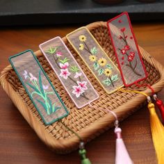 four embroidered bookmarks with tassels on a wicker tray next to flowers