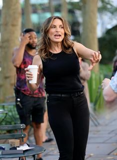 a woman holding a coffee cup in her right hand while standing next to a park bench