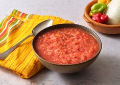 a bowl of salsa next to a small bowl of peppers on a yellow napkin with a spoon in it