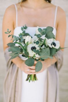 a woman holding a bouquet of flowers and greenery in her hands, wearing a white dress