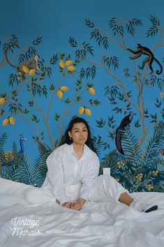 a woman sitting on a bed in front of a wall with birds and trees painted on it