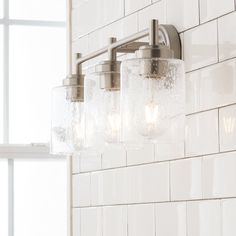 a bathroom light with three lights on it and white tile wall behind the light fixture