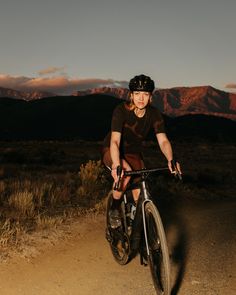 a woman riding a bike down a dirt road in the desert at sunset with mountains in the background