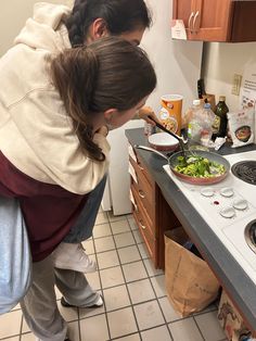 two women in the kitchen preparing food on the stove top burners, and one woman is bending over to pick something up with her hand