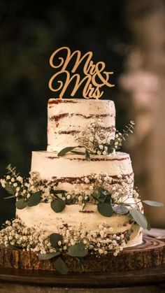 a wedding cake with greenery and mr and mrs topper on wooden slice stand