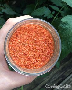 a hand holding a glass jar filled with red spices next to green leaves on a wooden table