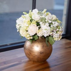 a gold vase filled with white flowers sitting on top of a wooden table next to a window