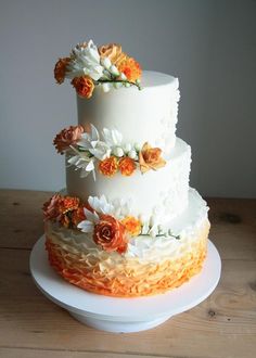 a three tiered white cake with orange and white flowers on the top, sitting on a wooden table